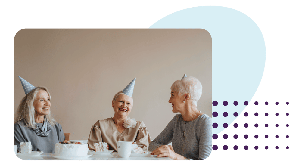 three senior citizen women celebrating birthday around a table. They are all wearing blue party hats and laughing.