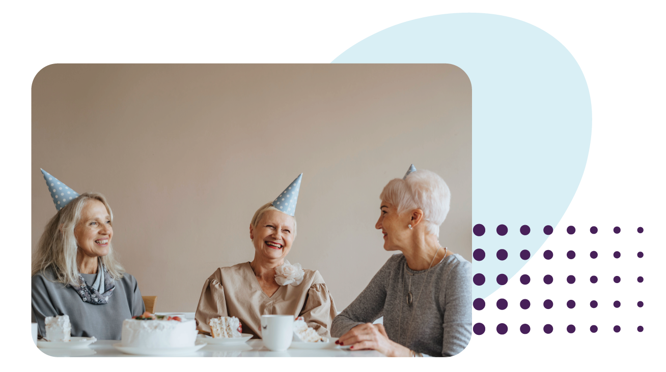 Three senior citizen women laughing around a table with blue party hats on.